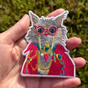 以下が含まれることがあります： 鮮やかなピンクの丸いメガネ、鮮やかなマゼンタのジャケット、青とピンクのアクセントが付いた重ね付けのゴールドネックレスを身に着けた、ふわふわした灰色の猫の刺繍ワッペン。猫は青い目を持ち、細部まで丁寧に表現されています。