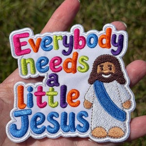 May include: An embroidered patch on a white background featuring the multi-colored text Everybody needs a little Jesus. To the right is a cartoon figure with brown hair, a white robe, a blue sash, and tan skin, all outlined in white thread.
