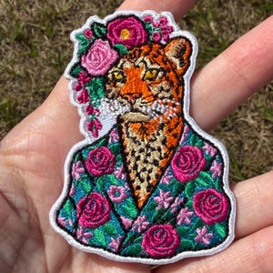 Könnte beinhalten: Ein gestickter Aufnäher mit einem Leoparden mit gelben Augen, der eine türkisfarbene Jacke mit einem rosa Rosenmuster trägt. Der Leopard hat eine Krone aus rosa und magentafarbenen Blumen auf dem Kopf. Der Aufnäher hat einen weißen Rand.