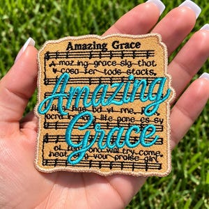May include: A rectangular embroidered patch featuring a tan background with black musical staff lines and lyrics. The text Amazing Grace is stitched in large, bright turquoise cursive script over the center of the black printed lyrics.