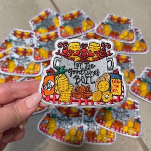 Op de afbeelding: Geborduurde patch met een pot rivierkreeften en de tekst "Let the good times BOIL". Het ontwerp bevat ananas, citroenen, hete saus en jam, geplaatst op een rood-wit geruit patroon.