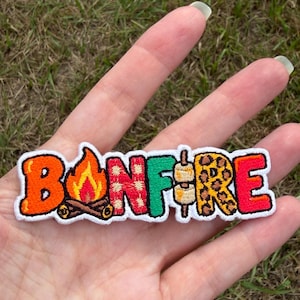 May include: An embroidered patch featuring the word BONFIRE in colourful, patterned letters. The B is orange, the O is a campfire, the N is red plaid, the F is green, the I is a marshmallow on a stick, the R is leopard print, and the E is red.