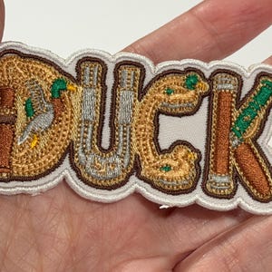 May include: An embroidered patch featuring the word DUCK in stylised lettering. The letters are filled with brown, tan, and silver thread, incorporating small green and grey duck motifs and brown shotgun barrel designs with a white border.