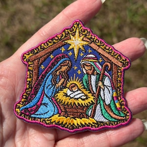 May include: An embroidered patch featuring a nativity scene with a central infant in a manger, flanked by two figures in blue and green robes, all set under a bright yellow star against a blue background with a dark pink border.