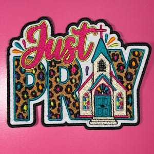 May include: An embroidered patch featuring the text Just PRAY in bright pink script and leopard print block letters. A small white church building with a teal roof, door, and stained glass windows is integrated into the design, outlined in black.