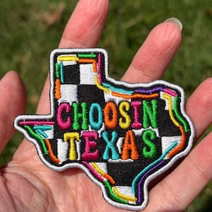 Könnte beinhalten: Ein bestickter Aufnäher in Form von Texas mit einem schwarz-weißen Schachbrettmuster als Hintergrund. Der Text "CHOOSIN TEXAS" ist in leuchtenden, mehrfarbigen Buchstaben gestickt. Der Rand ist weiß umrandet mit zusätzlichen neonfarbenen Akzenten.