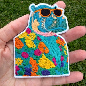 May include: An embroidered patch featuring a blue hippopotamus wearing orange-rimmed sunglasses. The hippo is dressed in a vibrant teal shirt covered in a colorful floral pattern of yellow, orange, pink, and purple flowers with green leaves.