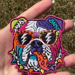 May include: An embroidered patch featuring a stylized bulldog head with a vibrant, multi-colored design. The dog wears sunglasses with lightning bolt details, has a spotted tongue hanging out, and wears a spiked collar in bright neon colors.