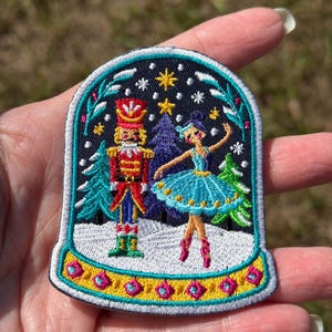 May include: An embroidered patch shaped like a snow globe featuring a nutcracker in a red uniform and a ballerina in a blue tutu standing in a snowy forest. The base is yellow with pink accents, and the dark sky is filled with stars.