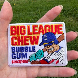 Puede incluir: Parche bordado con el texto "BIG LEAGUE CHEW BUBBLE GUM SINCE 1980" en rojo y azul. También presenta un jugador de béisbol con un bate. El parche es rectangular con fondo blanco.