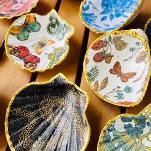 Op de afbeelding: Een verzameling decoratieve schelpvormige schaaltjes met goudkleurige randen. De schaaltjes hebben verschillende ontwerpen, waaronder vlinders, bloempatronen en een zwart-goudkleurig textuurpatroon. De schaaltjes zijn gerangschikt op een houten oppervlak.