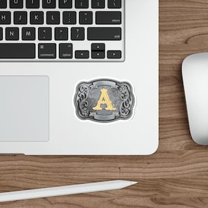May include: A sticker featuring a silver-colored, ornate belt buckle design with a gold letter "A" in the center. The buckle has a detailed floral pattern and is set against a white background.