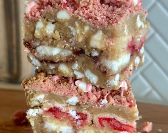 Puede incluir: Dos barras de postre apiladas con capas de chocolate blanco, fresas y una cobertura rosa desmenuzada. Las barras están sobre una superficie de madera, con migas esparcidas alrededor. El postre tiene una base marrón claro.