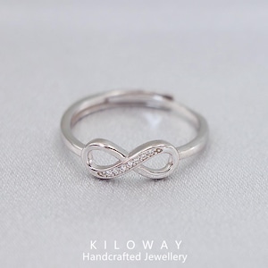 Könnte beinhalten: Verstellbarer Ring in Silber mit einem Unendlichkeitszeichen-Design. Das Unendlichkeitszeichen ist mit kleinen, klaren Edelsteinen besetzt. Der Ring wird auf einer hellgrauen Oberfläche gezeigt. Der Text "KILOWAY Handcrafted Jewellery" befindet sich unten.