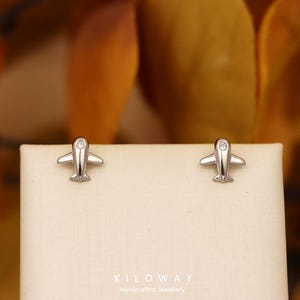 May include: Silver airplane stud earrings. Each earring has a small airplane design with a clear gemstone. The earrings are presented on a cream-coloured surface. The text "KILOWAY Handcrafted Jewellery" is visible at the bottom.
