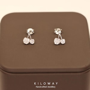 Puede incluir: Pendientes de botón de plata con forma de cereza. Cada pendiente presenta dos piedras redondas y brillantes, que se asemejan a cerezas, unidas por un fino tallo. Se exhiben sobre una superficie marrón. Se ve el nombre de la marca "KILOWAY Handcrafted Jewellery".