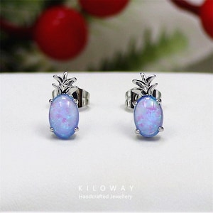 Puede incluir: Un par de pendientes de botón plateados con forma de piña. Cada pendiente presenta una piedra ovalada, opalina, de color azul claro con toques de rosa y morado. Los pendientes están sobre un fondo blanco. La marca "KILOWAY Handcrafted Jewellery" está impresa debajo.