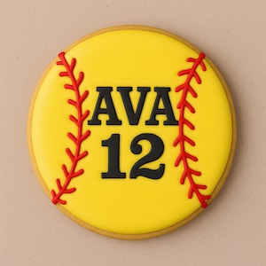 May include: A round, yellow cookie decorated to resemble a softball. The cookie features the name "AVA" and the number "12" in black, with red stitching details and a tan border.