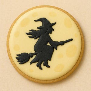 Puede incluir: Una galleta redonda de Halloween con una silueta negra de bruja montada en una escoba sobre un fondo de luna amarilla. La galleta tiene un borde marrón claro y está decorada con glaseado negro. El diseño está centrado en la galleta.