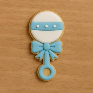 May include: A baby rattle cookie with a white circle top, a blue stripe with three white dots, and a blue bow. The handle and the outer edge of the cookie are light brown.