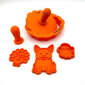 Op de afbeelding: Een oranje siliconen koekjessnijdersset met een pompoenvormige snijder, een kalkoen, een corgi en een spook. De pompoensnijder heeft een handvat en het woord "TOP" erop gedrukt. Ontworpen voor feestelijke traktaties.