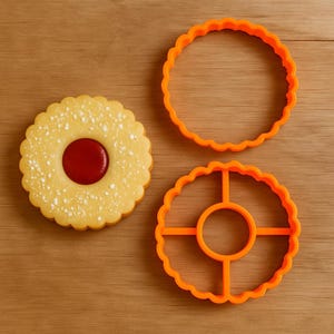 Könnte beinhalten: Nahaufnahme eines runden Kekses mit roter Mitte und zwei orangefarbenen Ausstechern. Der Keks hat einen gewellten Rand und ist mit Puderzucker bestäubt. Die Ausstecher sind ebenfalls rund mit gewellten Rändern.