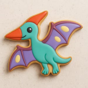 Peut inclure: Un biscuit décoré en forme de ptérodactyle. Le dinosaure a un corps turquoise, des ailes violettes avec des accents jaunes, et un bec et une crête orange. Le biscuit est bordé d'une couleur marron clair.