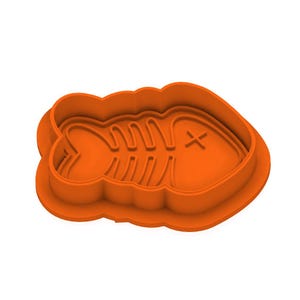 Puede incluir: Un cortador de galletas de plástico naranja con forma de esqueleto de pez. El cortador tiene un diseño detallado de los huesos y una X que marca el ojo. Diseñado para hacer galletas u otras golosinas.