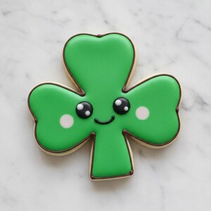May include: A green shamrock-shaped biscuit with a smiling face, black eyes, and white cheeks. It has a brown outline and a pale base. The image is a close-up shot.