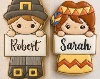 May include: Two decorated cookies in the shape of cartoon figures. One is a pilgrim with a hat and the name "Robert" on a sign. The other is a Native American girl with the name "Sarah" on a sign. The cookies are decorated with icing.