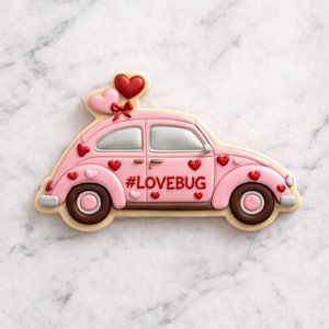 Op de afbeelding: Een roze koekje in de vorm van een auto, versierd met rode hartjes en de tekst "#LOVEBUG". Het koekje heeft bruine wielen en een hartvormige ballon erbovenop. Een traktatie met Valentijnsthema.