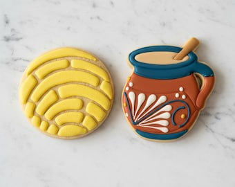 May include: Two decorated sugar cookies. One is a round yellow cookie with a raised shell-like pattern. The other is a brown clay-style mug with a blue rim and handle, containing a tan liquid and a small wooden stirring stick, featuring white details.