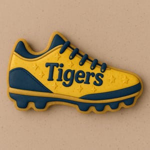 May include: A yellow and navy blue football cleat-shaped cookie with the word "Tigers" in navy blue. The cookie has star-shaped sprinkles and a navy blue trim. The laces are navy blue.
