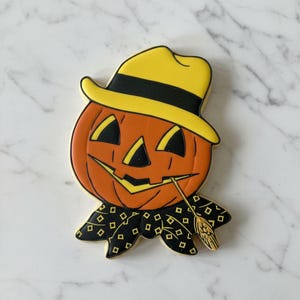 May include: A pumpkin-shaped cookie decorated with orange icing, featuring a yellow hat with a black band, black triangular eyes and nose, and a wide grin. A black scarf with a yellow diamond pattern is around the neck, with a piece of wheat.