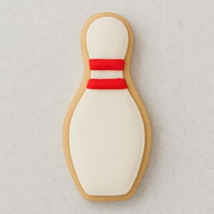 Puede incluir: Una galleta con forma de bolos con un centro blanco y dos rayas rojas. La galleta está delineada en un color marrón claro, que se asemeja a un diseño clásico de bolos. Ideal para un evento deportivo.