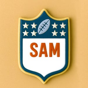 May include: A shield-shaped cookie with a blue border and a football design at the top. The cookie has white stars and the name "SAM" in orange letters. The background is a solid yellow color.