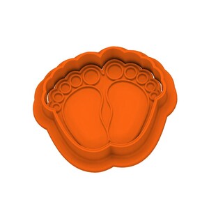 Puede incluir: Un cortador de galletas de plástico naranja con forma de dos pies de bebé. El cortador tiene un borde festoneado e impresiones detalladas de los dedos de los pies y las plantas de los pies. El diseño es una forma sólida tridimensional.
