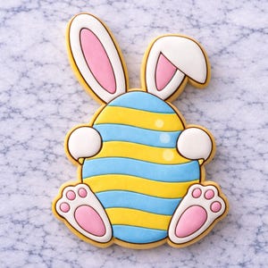 Easter bunny egg sugar cookie decorated with royal icing, featuring a bunny peeking from behind a blue and yellow striped Easter egg on a marble background