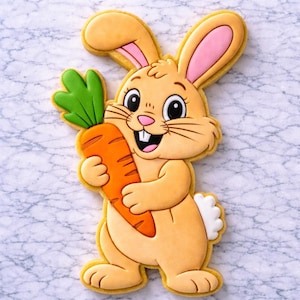 Könnte beinhalten: Ein dekorierter Keks in Form eines Cartoon-Hasen, der eine Karotte hält. Der Hase ist hellbraun mit rosa Innenohren und einem weißen Schwanz. Die Karotte ist orange mit grünen Blättern.