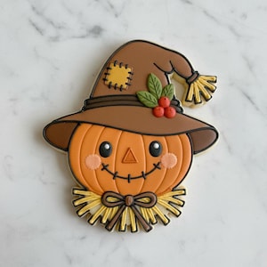 May include: A decorated sugar cookie shaped like a scarecrow with a round orange pumpkin face, a brown hat with a yellow patch, green leaves, and red berries. The scarecrow features a black stitched smile, pink cheeks, and yellow straw accents.