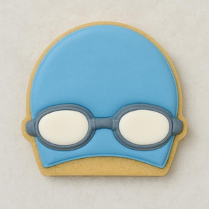 Può includere: Un biscotto azzurro a forma di cuffia e occhialini da nuoto. La cuffia è di un azzurro uniforme, e gli occhialini sono grigi con lenti bianche. Il biscotto ha un bordo marrone chiaro.