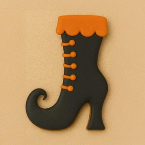Puede incluir: Una galleta con forma de bota de bruja negra con detalles en naranja. La bota tiene una punta curvada, un tacón alto y botones naranjas. La parte superior de la bota tiene un borde naranja festoneado. La galleta está sobre un fondo beige.