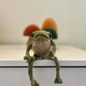 May include: A handmade, knitted frog doll in shades of green and tan, with black eyes, sits on a white surface. The frog's long legs dangle over the edge. In the background, there are three decorative mushroom figures.
