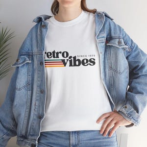 May include: White t-shirt with the text "retro. Vibes" in black, with a retro color stripe design. The shirt also includes the text "SINCE 1975". The model is wearing a light blue denim jacket.