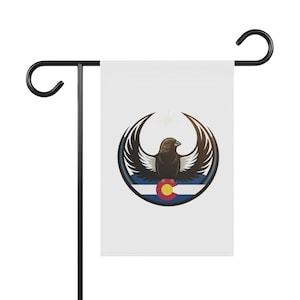 Op de afbeelding: Een witte tuinvaandel met een gestileerde vogel met uitgespreide vleugels, tegen een blauw-witte gestreepte achtergrond. Het embleem bevat de vlag van de staat Colorado. De vlag is gemonteerd op een zwarte metalen standaard.