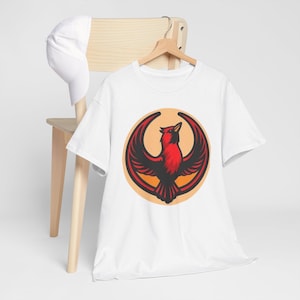 May include: White t-shirt featuring a red and black bird design with outstretched wings, set against an orange circle. The bird is the focal point, with a detailed illustration. A white baseball cap rests on a wooden chair.