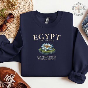 May include: Navy blue sweatshirt with the word "EGYPT" in a cream-colored font. Below the word "EGYPT" is the text "GIFT OF NILE" and an illustration of an Egyptian lotus flower. The text "EGYPTIAN LOTUS Nymphaea caerulea" is below the illustration.