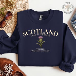 May include: Navy blue sweatshirt with the word "SCOTLAND" in cream-colored letters. Below the word "SCOTLAND" is the phrase "LAND OF THE BRAVE" and a thistle illustration. The word "THISTLE" and the scientific name "Onopordum acanthium" are below the illustration.