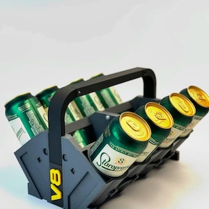 May include: A gray and black beer caddy with a handle, holding multiple green and gold beer cans. The caddy has the yellow letters "V8" on the side. The beer cans have the text "Staropramen" on them.