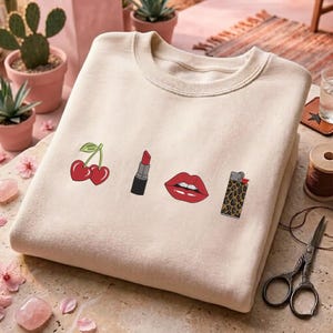 May include: Cream-colored sweatshirt featuring embroidered designs of a pair of cherries, a lipstick, a pair of red lips, and a leopard print lighter. The sweatshirt is folded and displayed on a stone surface.